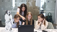 Jugendliche schauen auf einen Bildschirm, sie programmieren den humanoiden Roboter NAO unter Anleitung der Lehrerin, die erklärend hinter ihnen steht.